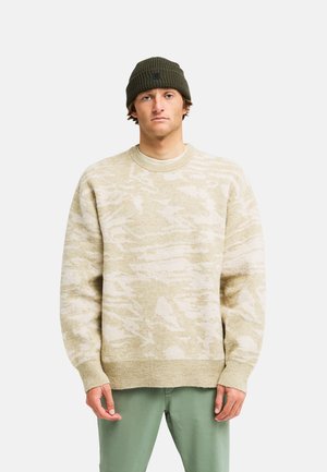 Jonge man draagt een beige trui met camouflagepatroon, een olijfgroene muts en lichtgroene broek, staand tegen een effen achtergrond.