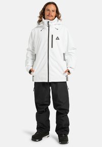 Witte waterdichte jas met een capuchon, zwarte accenten en een borstzak. Gepaard met zwarte cargo-broeken en zwarte laarzen.