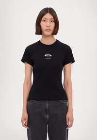 Kvinna med rakt axellångt hår bär en svart Moschino Jeans-t-shirt och mörka denimjeans, stående mot en helt vit bakgrund.