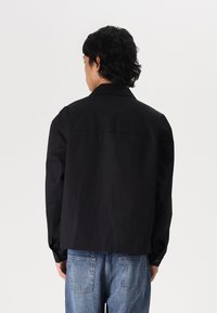 Črna jakna s ovratnikom, dolgimi rokavi in gumbi. Oblečena čez svetlo modre jeans hlače, ki prikazujejo enobarven dizajn in gladko teksturo.