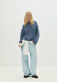 Blauer gestrickter Pullover mit einer Tasche, kombiniert mit hellblauen, gestreiften, weiten Hosen und braunen Slippern. Weißer Stoff um die Taille gebunden.