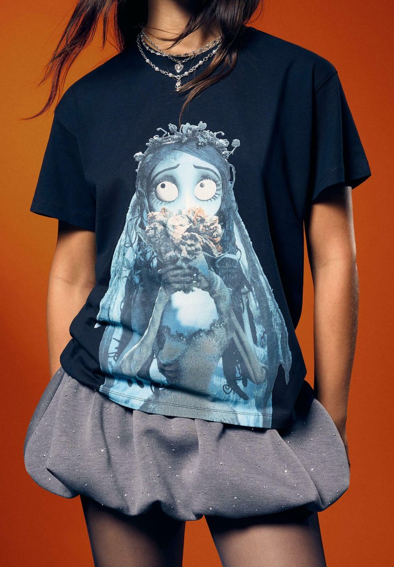 Schwarzes T-Shirt mit einer Illustration eines blauhäutigen Charakters mit langen Haaren, der Blumen hält. Graue, strukturierte Shorts mit einem Bund-Detail.