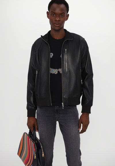 Μαύρο δερμάτινο τζάκετ bomber με φερμουάρ και ριχτές μανσέτες, φορεμένο πάνω από μαύρο πουκάμισο, συνδυασμένο με σκούρους γκρι τζιν και μια ριγέ τσάντα.