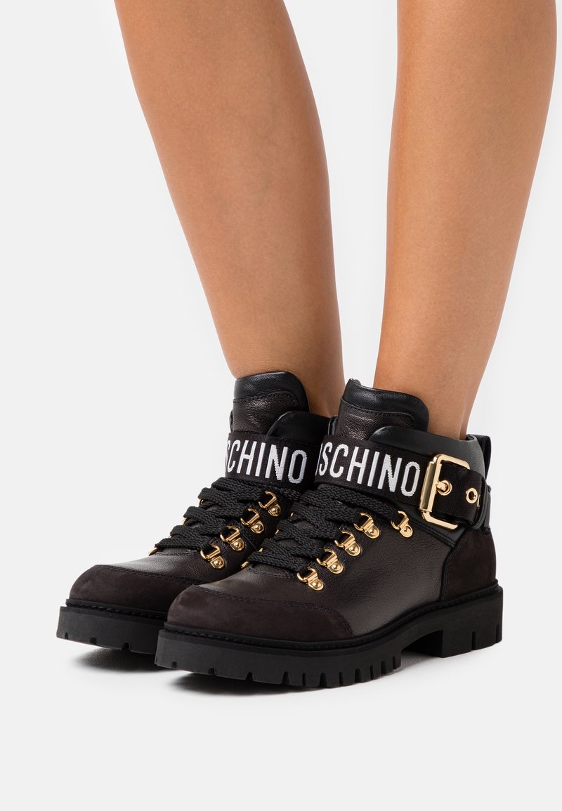 Bottines en cuir noir avec semelles épaisses, crochets à lacets dorés, lacets larges, bride à boucle dorée et bande blanche avec le texte "MOSCHINO" autour de la cheville.