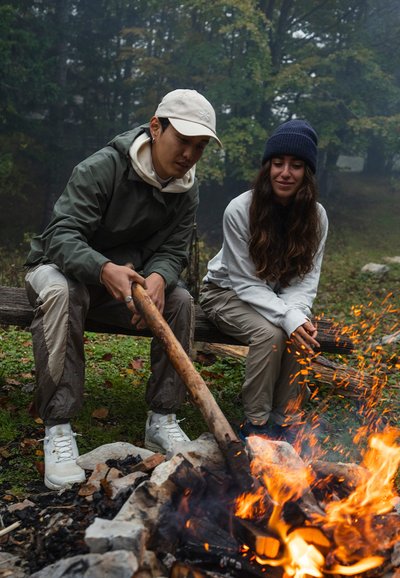 Dva lidé sedí u táboráku. Jeden drží klacek; oba jsou oblečeni v outdoorovém oblečení. Scéna obsahuje oranžové plameny a kouř, obklopené zelení.