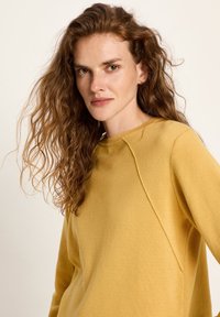 Frau mit langen, lockigen roten Haaren, die ein senfgelbnes Langarmoberteil trägt und direkt in die Kamera schaut, vor einem schlichten Hintergrund.