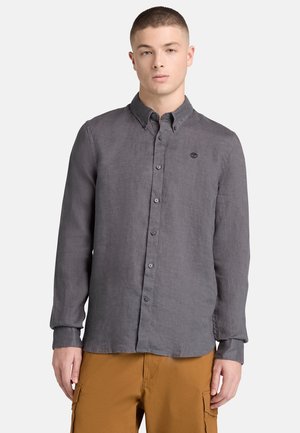 Junger Mann mit einem dunkelgrauen, langärmligen Hemd mit Knöpfen und senffarbenen Cargohosen, der vor einem schlichten, hellen Hintergrund steht.