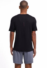 T-shirt de sport noir à manches courtes avec un discret accent vertical rouge dans le dos, texture lisse, associé à un short gris clair.