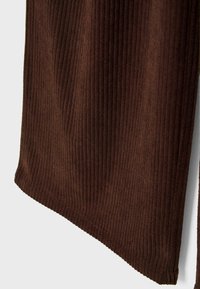 Gros plan d'un rideau en tissu marron côtelé suspendu devant un fond blanc avec une texture verticale douce et un ourlet cousu.