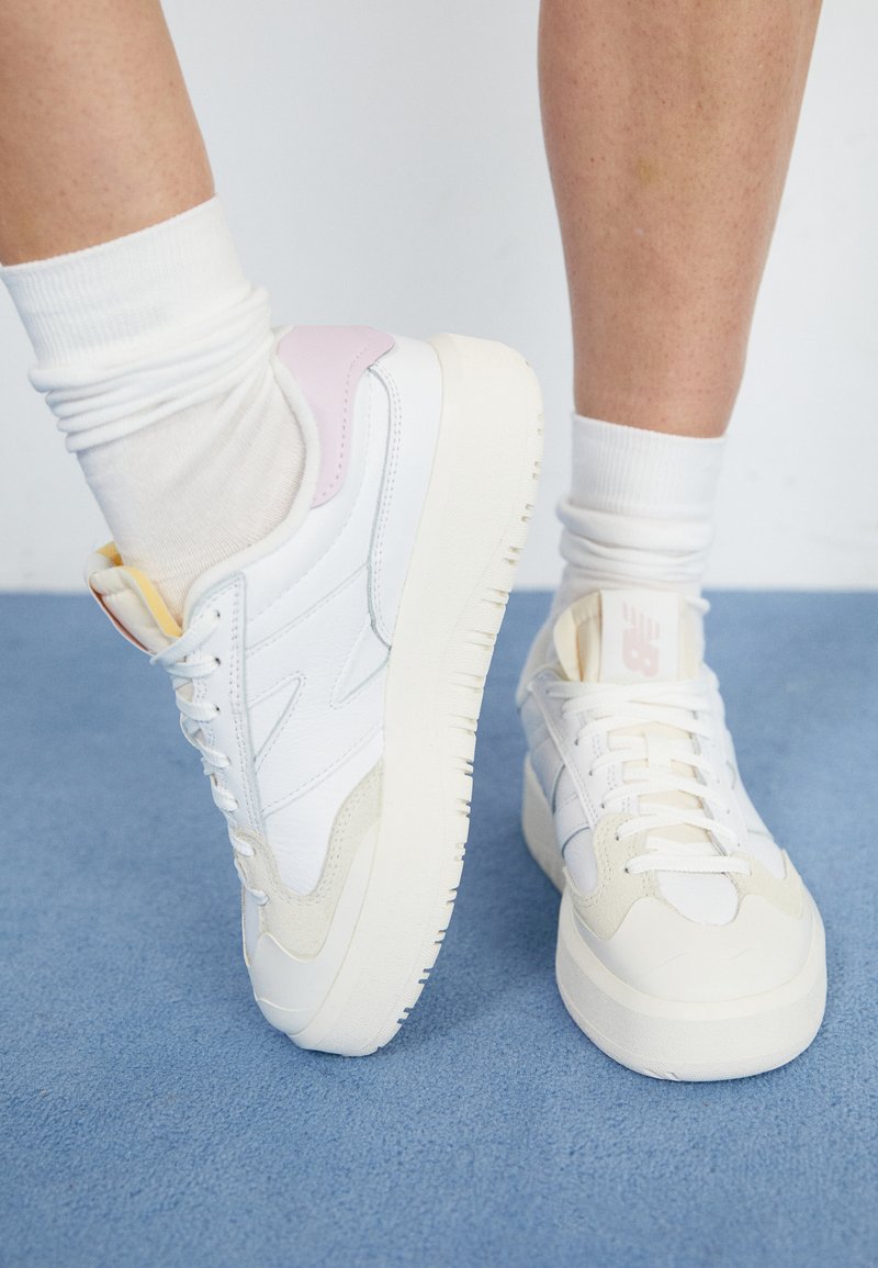 Des baskets blanches avec une semelle épaisse, ornées d'accents pastel rose et jaune, fabriquées en cuir lisse et en matériaux synthétiques, avec des fermetures à lacets.