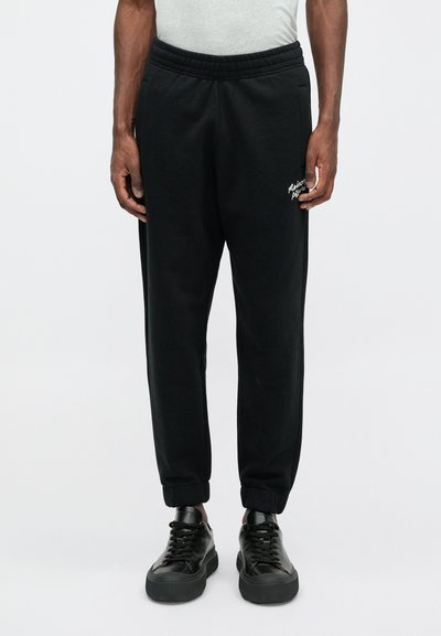 Pantaloni de trening negri, confecționați dintr-un material moale, cu talie elastică și mansete, având un logo alb pe șoldul stâng. Purtați cu adidași negri.
