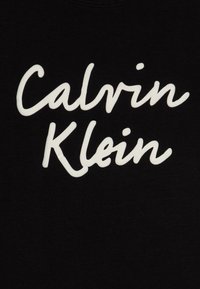 Czarna koszulka z białym, kursywnym napisem „Calvin Klein”. Tkanina wygląda na gładką i ma klasyczny okrągły dekolt.