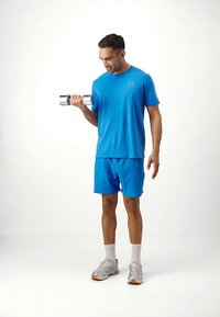 Άνδρας με μπλε αθλητική στολή σηκώνει ασημένιο αλτήρα με το δεξί του χέρι, στέκεται σε λευκό δάπεδο μπροστά από απλό λευκό φόντο.