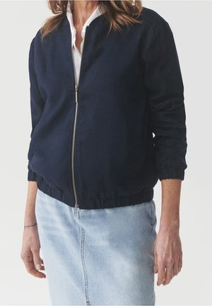 Kobieta w granatowej kurtce na zamek błyskawiczny nałożonej na białą koszulę, w połączeniu z jasnoniebieską spódnicą dżinsową, stoi na jednolitym tle.