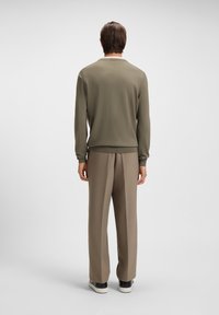 Mann, der mit dem Rücken zur Kamera steht, trägt einen olivgrünen Pullover, taupefarbene Hosen und schwarz-weiße Sneakers vor einem schlichten hellen Hintergrund.