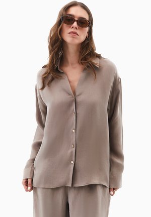 Femme portant une chemise taupe ample boutonnée et un pantalon assorti, accessoirisée de lunettes de soleil écaille et de boucles d’oreilles créoles en or.
