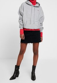 Un sweat à capuche gris avec une poche avant, superposé sur un col roulé rouge, associé à une jupe en denim noire et des bottines noires.