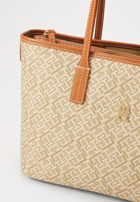 Beige stoff tote bag med et geometrisk mønster, brune skinndetaljer og en gyllen logo, med en topp glidelåslukking og to skulderstropper.