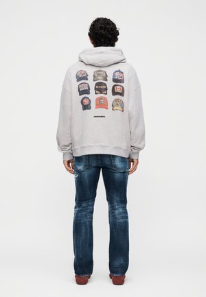 Άνδρας με σγουρά μαλλιά που φοράει ανοιχτογκρι φούτερ με κουκούλα και εικόνα εννέα καπέλων μπέιζμπολ και μπλε τζιν, στέκεται με την πλάτη προς τα εμπρός σε λευκό φόντο.