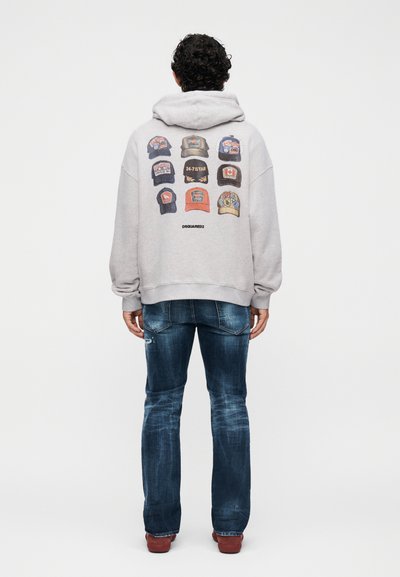 Hombre con cabello rizado que lleva una sudadera con capucha gris claro con un gráfico de nueve gorras de béisbol y vaqueros azules, de pie de espaldas sobre un fondo blanco.