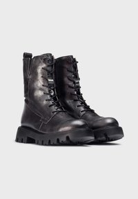 Botas de tobillo de cuero negro con un acabado mate, puntera reforzada y suela de goma resistente; cuenta con cordones en la parte delantera y una lengüeta en la parte trasera.