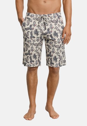 Mann trägt beige kniehohe Shorts mit blauem und rotem Paisley-Muster und Kordelzug in der Taille, steht barfuß mit den Händen in den Taschen.