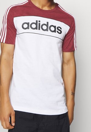 Osoba nosząca koszulkę Adidas z krótkim rękawem, bordowymi ramionami i rękawami, białym tułowiem oraz czarnym logo na piersi.