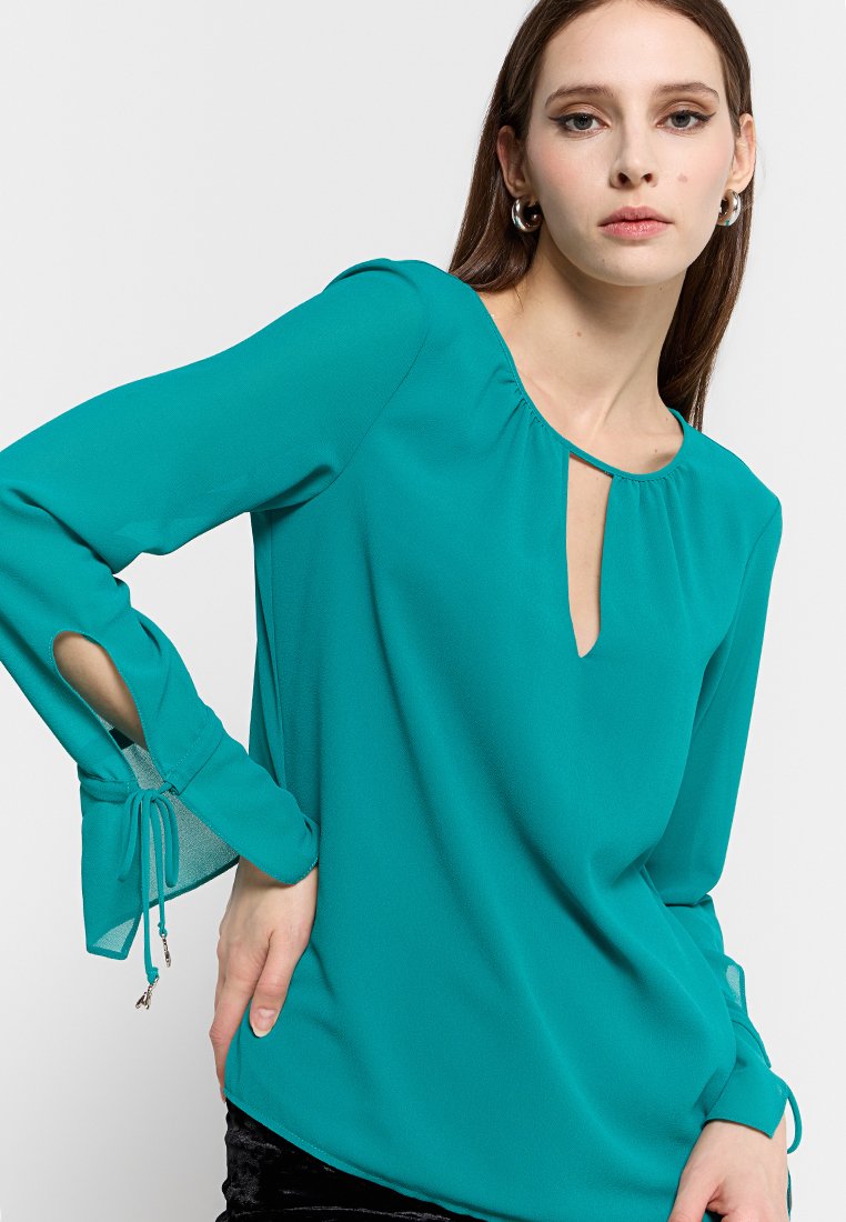 Türkise Bluse mit rundem Ausschnitt, Schlüssel-Ausschnitt-Detail und langen Ärmeln. Schlitze an den Ärmeln mit Bindebändern. Glattes, leichtes Material.