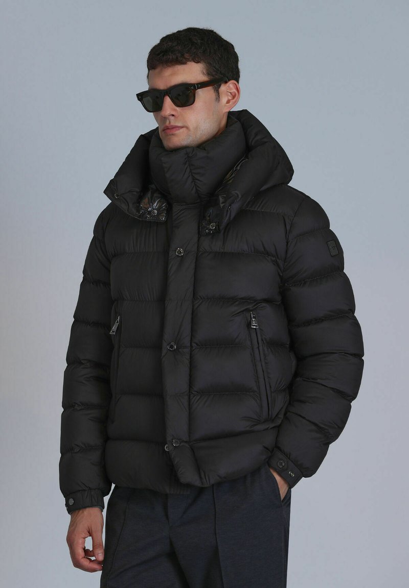 Black puffer jacket with a high collar, glossy accents, two front zip pockets, and a lightweight, quilted design. Worn with sunglasses.