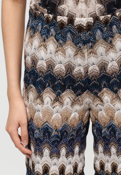 Κοντινό σε χέρι και μπράτσο ενός ατόμου δίπλα σε παντελόνι με υφασμένο ζιγκ ζαγκ μοτίβο σε αποχρώσεις μπλε, καφέ, μπεζ και λευκού.