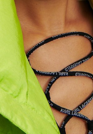 Gros plan sur le haut de la poitrine d'une personne avec des lacets noirs croisés sur la peau et un vêtement vert vif sur le côté gauche.