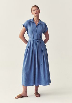 Kvinde iført en midt-længde blå knapkjole med et bundet bælte i taljen og korte ærmer, stående med hænderne bag ryggen og brune slip-in sandaler.