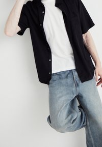 Camisa negra de manga corta con botones sobre una camiseta blanca, combinada con unos vaqueros ligeros de corte holgado en azul claro, destacando la superposición casual y la textura.