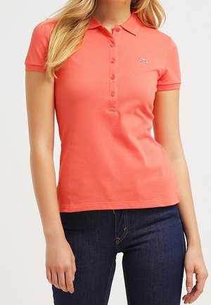 Kobieta w koralowo-różowej koszulce polo z krótkim rękawem i guziczkami oraz ciemnoniebieskich dżinsach, stojąca na jednolitym tle.