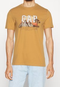 T-shirt en coton jaune moutarde présentant un motif de montagnes et de palmier avec le lettrage "GAP" en blanc et gris. Manches courtes, col rond.