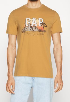 T-shirt en coton jaune moutarde présentant un motif de montagnes et de palmier avec le lettrage "GAP" en blanc et gris. Manches courtes, col rond.