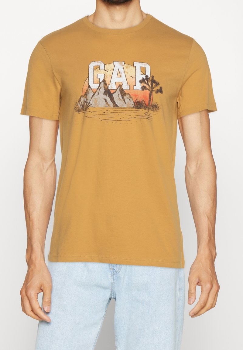 T-shirt en coton jaune moutarde présentant un motif de montagnes et de palmier avec le lettrage "GAP" en blanc et gris. Manches courtes, col rond.