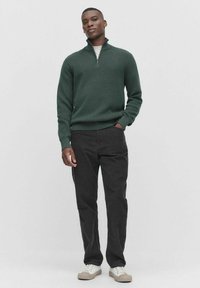 Ciemnozielony sweter z prążkowanym wzorem i półzamkiem, w zestawieniu z czarnymi spodniami i jasnymi sneakersami. Model stoi na tle jednolitego tła.