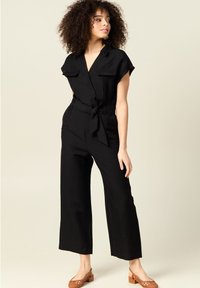 Svart linjumpdräkt med korta ärmar, krage, framfickor och knytbart midjebälte. Återfinns med bruna vävda blockklacksskor.