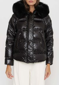 Giacca a vento nera con finitura lucida, cappuccio foderato in pelliccia, sezioni trapuntate, due tasche frontali e polsini elasticizzati.