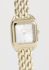 Montre-bracelet dorée avec un boîtier carré, un cadran blanc, des aiguilles dorées et des index en chiffres romains. Équipée d'un bracelet en métal poli.