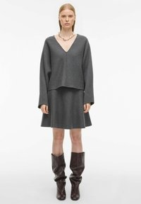 Šedý top z vlněné směsi s výstřihem do V a dlouhými rukávy, v kombinaci s odpovídající rozšířenou sukní. Modelka má na sobě hnědé kožené boty do půli lýtek.