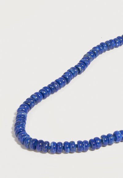 Sautoir de perles rondes bleues polies avec de légères variations de couleur et de texture, disposé en courbe lâche sur fond blanc.