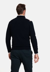 Granatowy sweter z dzianiny z teksturowanymi akcentami na ramionach, ściągaczowymi mankietami i dołem oraz klasycznym kołnierzykiem, w zestawie z ciemnozielonymi spodniami.