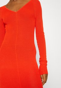 Robe rouge côtelée à manches longues et col en V. Présente une silhouette ajustée et un motif en tricot texturé, mettant en valeur une finition lisse.