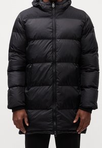 Veste d'hiver noire matelassée avec fermeture éclair avant et poches latérales zippées, portée par une personne debout, visible des épaules aux cuisses.