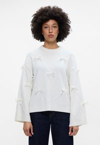 Balta megztinė su deimanto raštu ir tekstūruotais kaspinais. Savaitiniai rankovės ir banguota apykaklė. Minkšta, pūkuota medžiaga.