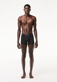 Fekete boxernadrág elasztikus derékpánttal, melyen a "LACOSTE" felirat látható. Sima anyag, slim fit dizájn, és combközép hosszúság. Mezítláb, álló helyzetben.