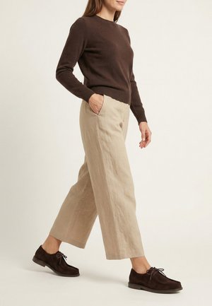 Frau läuft, trägt einen dunkelbraunen Pullover, beige weite Hosen und dunkelbraune Schnürschuhe vor hellem Hintergrund.