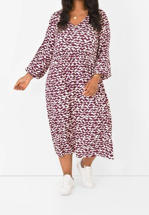 Vrouw draagt een paars-witte midi-jurk met lange mouwen en witte sneakers tegen een effen witte achtergrond.